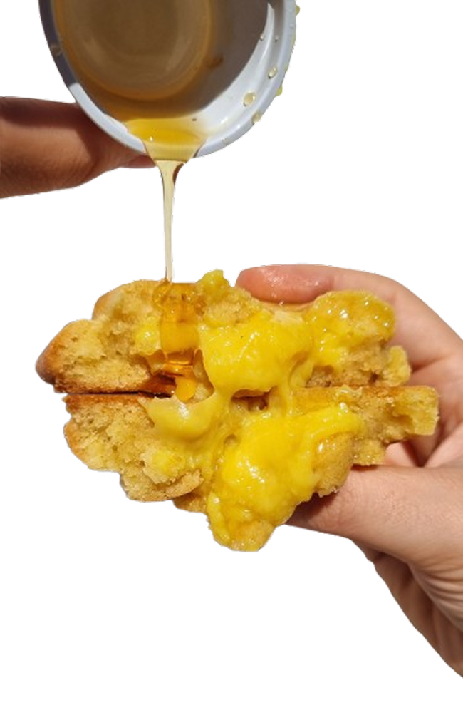 
A imagem exibe um pedaço de cookie cortado ao meio, revelando um recheio abundante de cor amarela vibrante e textura cremosa. Uma mão está segurando a parte de baixo do cookie, enquanto outra mão, no canto superior esquerdo, derrama um líquido dourado e viscoso, que parece é mel, sobre o recheio exposto. O recheio amarelo escorre um pouco, dando a impressão de ser bem macio e talvez quente. O fundo da imagem é dividido horizontalmente: a parte superior é de um tom de amarelo mostarda sólido, enquanto a parte inferior é branca. A cena sugere um alimento doce, indulgente e reconfortante.